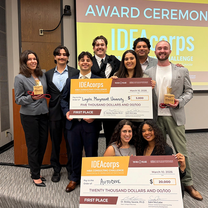 The IDEACorps MBA Consulting Challenge 2026 Team with their faculty advisor and client holding up two large checks.